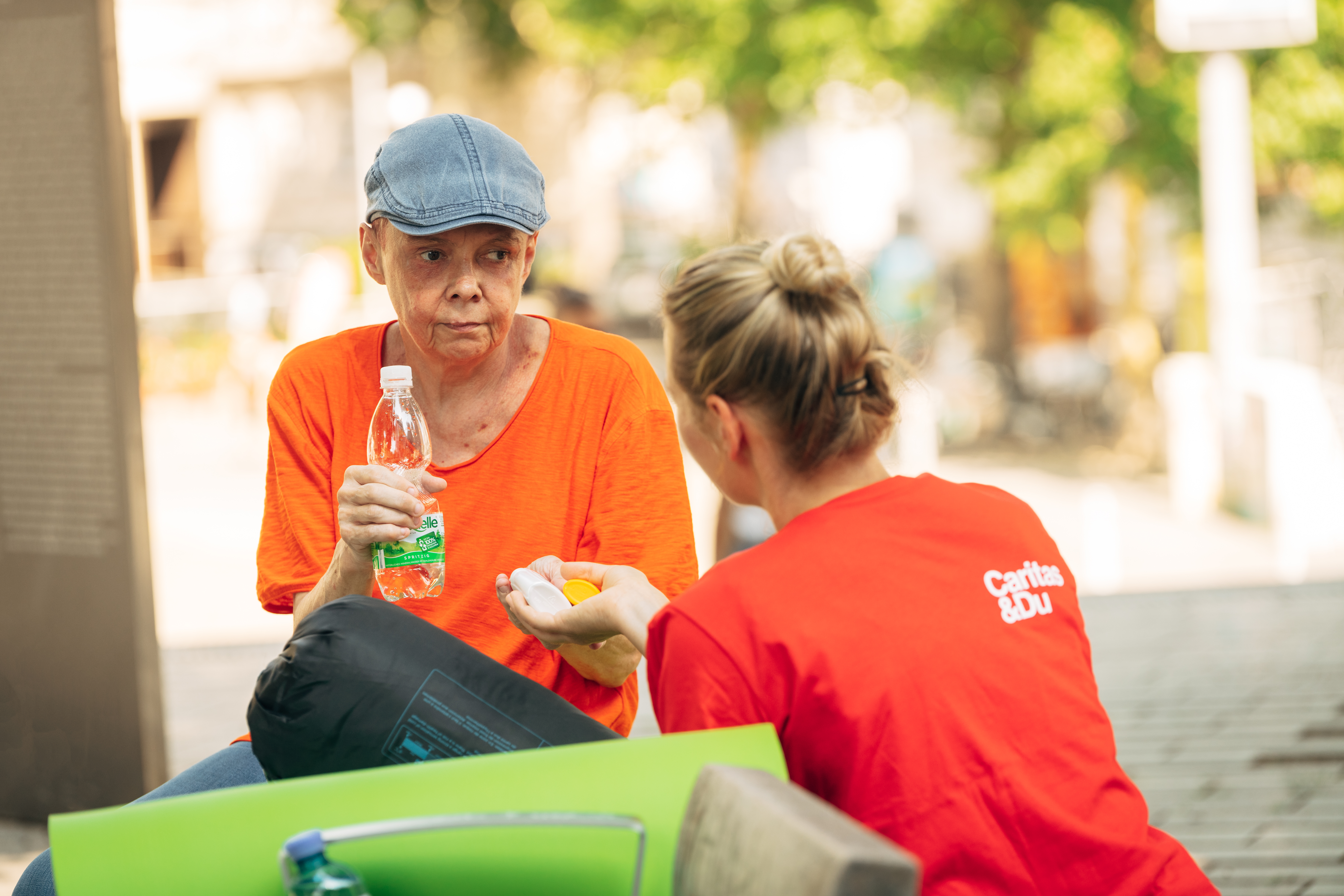 Eine Caritasmitarbeiterin reicht einer obdachlosen Person Wasser und Sonnencreme
